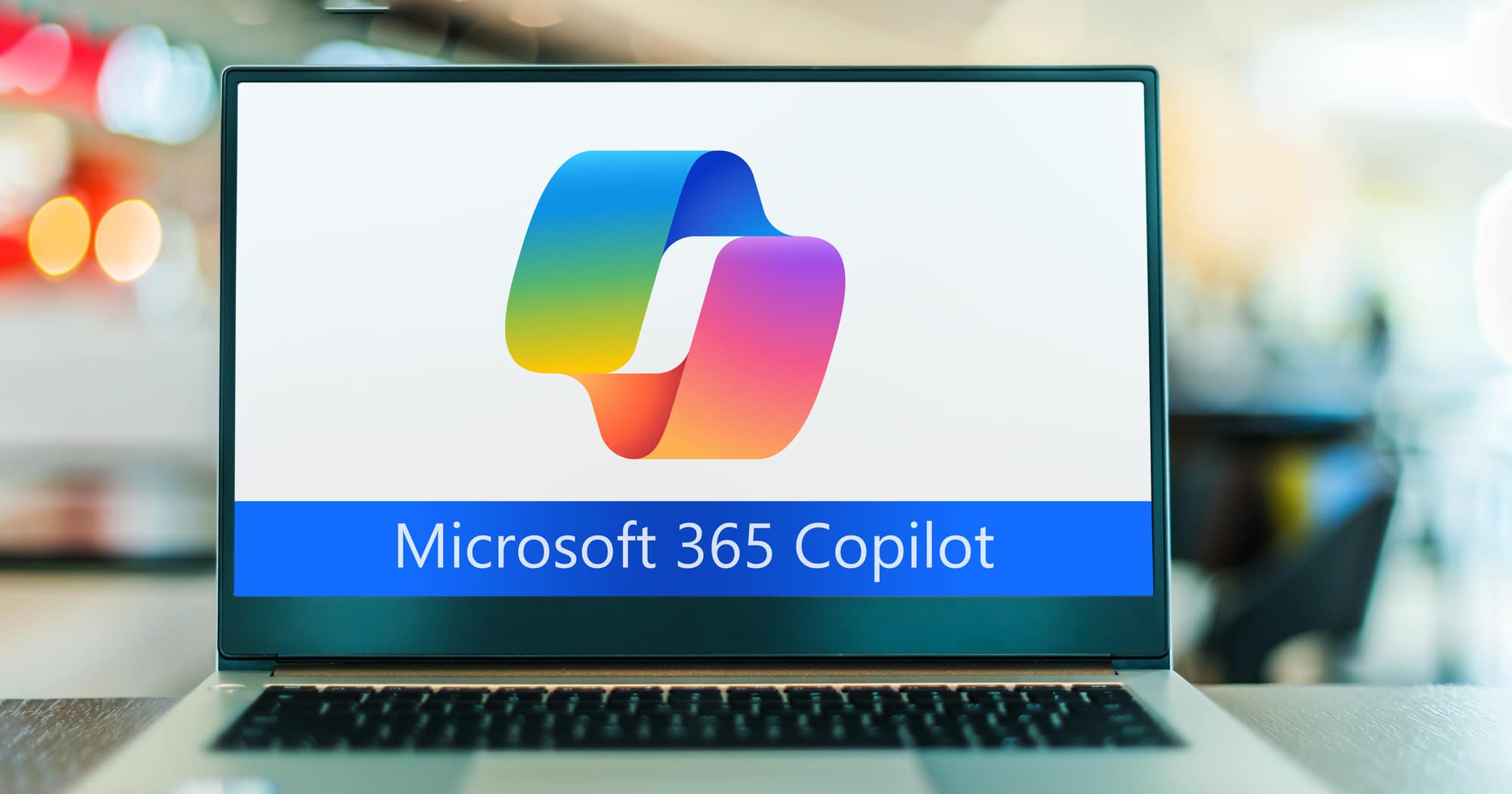 A laptop screen displaying the logo of Microsoft 365 Copilot pro. The logo consists of a multicolored shape resembling a folded ribbon in blue, green, yellow, and pink hues, positioned above the text "Microsoft 365 Copilot" in blue font. The laptop is on a table with a blurred background, suggesting a busy office or public space setting.