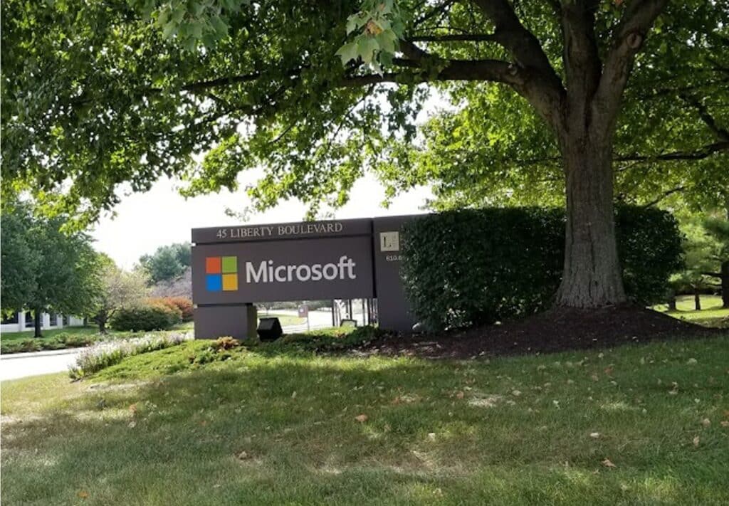 
A serene outdoor scene at the entrance of a Microsoft campus located at 45 Liberty Boulevard. The company's name is prominently displayed on a modern, dark signboard with the iconic Microsoft logo in its colorful square design. Lush green trees and well-maintained landscaping surround the sign, underlining the tech giant's presence in a peaceful, natural setting. This is a place where innovations such as Copilot Pro are developed, contributing to Microsoft's legacy of technological advancement and productivity solutions.