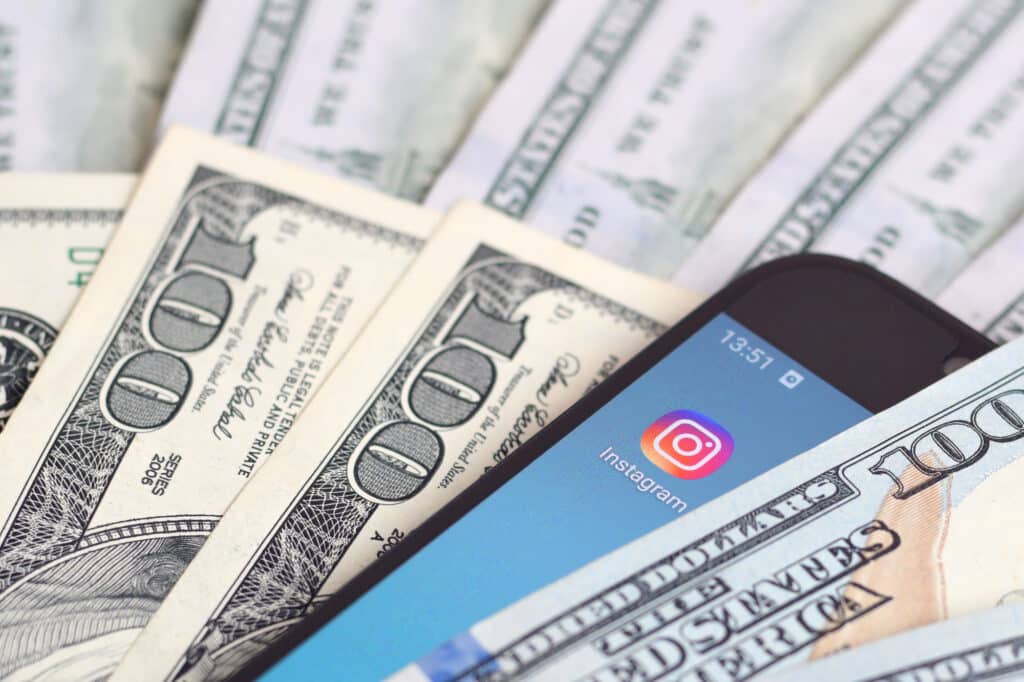 An image showing a smartphone displaying the Instagram app, surrounded by US hundred-dollar bills, representing the lucrative nature of an AI Instagram Influencer.