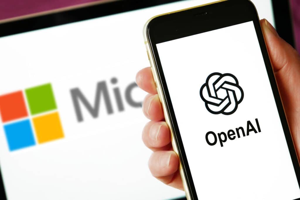 A hand showcasing the ChatGPT vs. Copilot rivalry with a smartphone displaying the OpenAI logo and a Microsoft logo in the background.