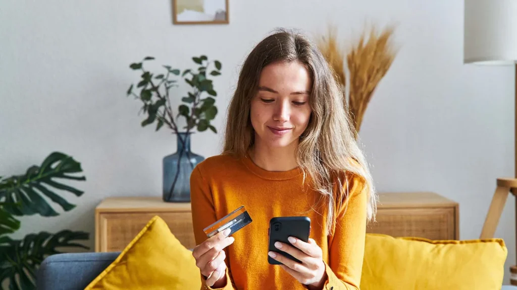 Woman shopping online with a phone and payment card, showing how Intelligent Commerce can connect AI-assisted product choices with secure checkout.